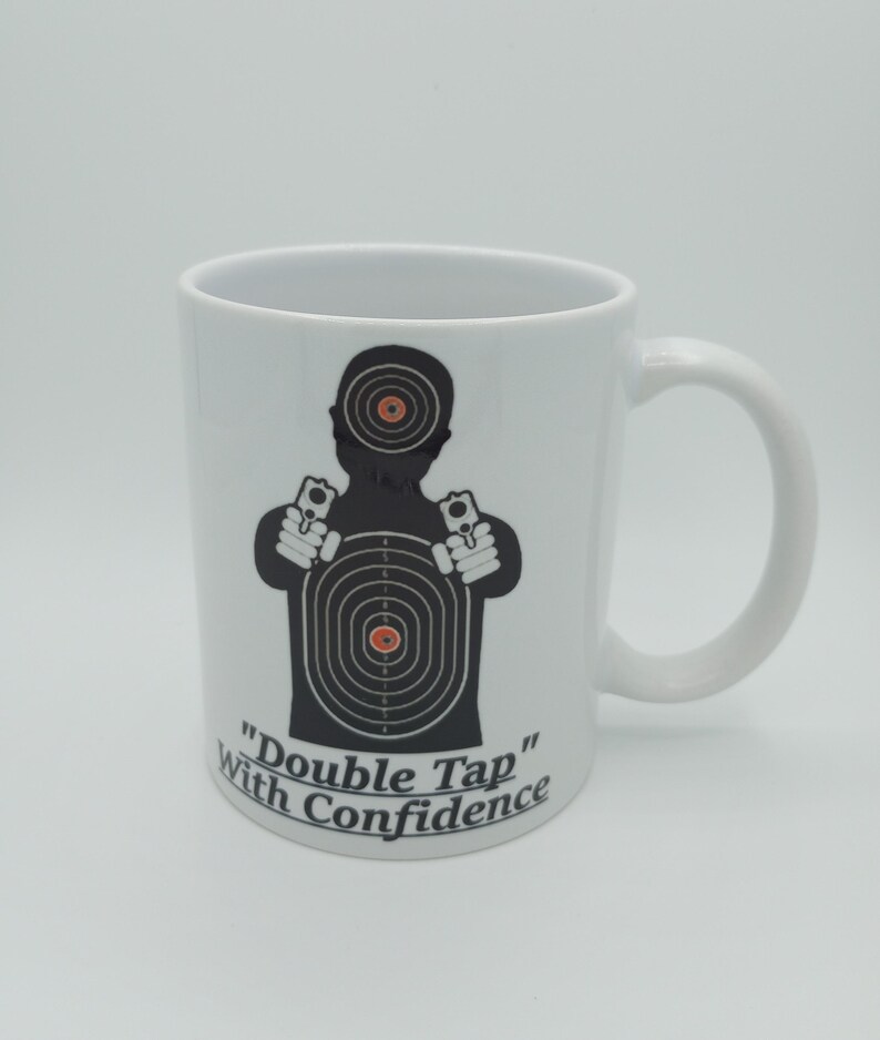 K&ouml;nnte beinhalten: Eine wei&szlig;e Keramiktasse mit einer schwarzen Silhouette einer Person, die zwei Pistolen vor einer Zielscheibe h&auml;lt. Der Text "Double Tap" und "With Confidence" ist unter der Silhouette aufgedruckt.