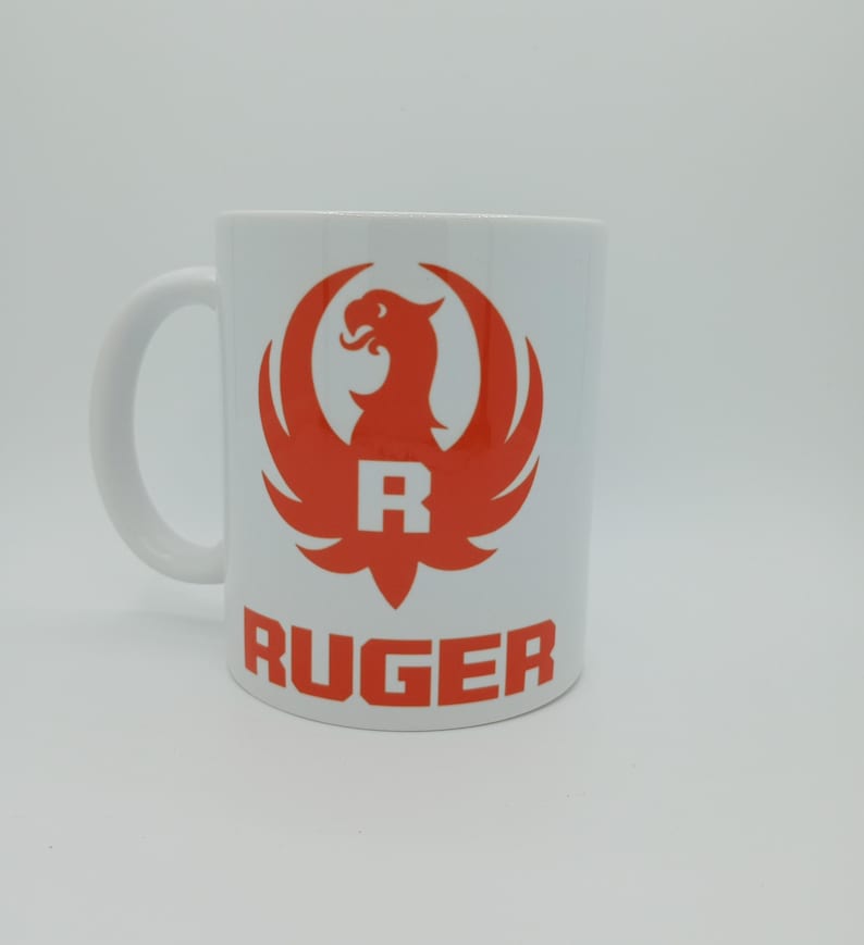 K&ouml;nnte beinhalten: Wei&szlig;e Keramiktasse mit einem roten Ruger-Logo, das einen stilisierten Vogel mit ausgebreiteten Fl&uuml;geln und dem Buchstaben "R" in der Mitte zeigt. Das Wort "RUGER" ist in Rot unter dem Logo gedruckt.