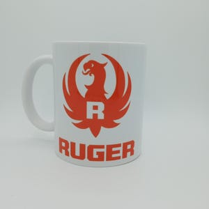 K&ouml;nnte beinhalten: Eine wei&szlig;e Keramiktasse mit einem roten Ruger-Logo, das einen stilisierten Vogel mit ausgebreiteten Fl&uuml;geln und dem Buchstaben "R" in der Mitte zeigt. Das Wort "RUGER" ist in Rot unter dem Logo gedruckt.