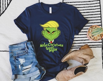 May include: Navy blue t-shirt featuring a green Grinch character with yellow hair, a red tie, and the phrase "Make Christmas Great Again." The shirt is paired with denim shorts and tan ankle boots, set against a patterned rug.
