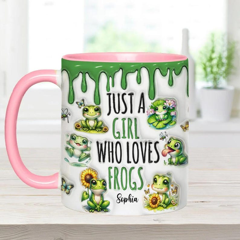 May include: A white ceramic mug with a pink rim and green dripping paint design. It features a cartoon frog design with the text "Just a girl who loves frogs" and a personalized name "Sophia".