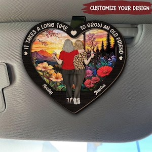 May include: A heart-shaped wooden car charm with a stained glass design featuring two women standing in a field of flowers. The text reads "It takes a long time to grow an old friend." The names "Ansley" and "Jessica" are written at the bottom of the charm.