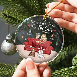 May include: A round, clear glass ornament with a red, white, and black illustration of a couple sitting on a red couch with two cats. The ornament has the text "I love you as much as the cats (almost)" and the names "Ethan" and "Anne".