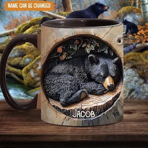 May include: A brown ceramic mug featuring a black bear sleeping in a tree trunk design. The mug has a black handle and the name "Jacob" is printed on the front.