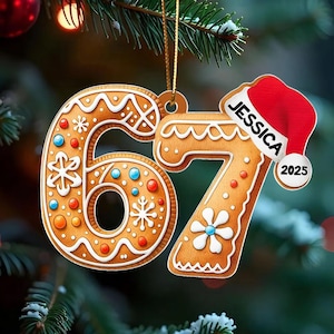 May include: Gingerbread-style Christmas ornament shaped like the numbers 67, decorated with white icing and colorful sprinkles. A small Santa hat with the name "JESSICA" and the year "2025" is attached. The ornament hangs from a gold string.
