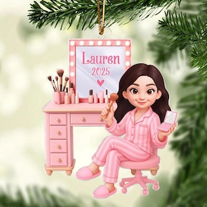 May include: A pink Christmas tree ornament depicting a cartoon girl in striped pyjamas at a dressing table. The mirror reads "Lauren 2025". Makeup brushes and products are on the table. The girl holds a makeup brush and compact.