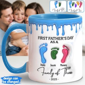 Op de afbeelding: Een witte keramische mok met een blauwe rand en blauwe verf die langs de zijkant druipt. De mok heeft een ontwerp met drie voetafdrukken met de tekst "First Father's Day as a Family of Three - 2025" erop gedrukt.