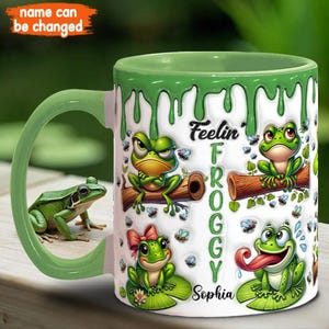 May include: A white ceramic mug with a green rim and a green drip design. The mug features a cartoon frog design with the text "Feelin' Froggy Sophia".