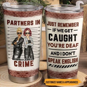 Puede incluir: Dos tazas de viaje de acero inoxidable con texto e ilustraciones humorísticas. Una taza dice "Partners in Crime" con dos mujeres con gafas de sol. La otra taza dice "Just Remember - If We Get Caught You're Deaf And I Don't Speak English".