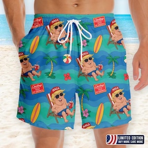 Op de afbeelding: Blauwe zwembroek met een herhalend patroon van een stripfiguur met een zonnebril en een "MAGA"-hoed, liggend in een strandstoel. De zwembroek heeft ook surfplanken, palmbomen en de tekst "Greetings Gulf of America".