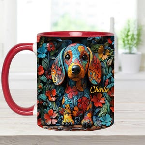 Puede incluir: Una taza de caf&eacute; de cer&aacute;mica roja y blanca con una ilustraci&oacute;n colorida de un perro salchicha con patrones florales. El nombre del perro, "Charlie", est&aacute; escrito en texto amarillo en la taza.