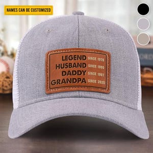 Könnte beinhalten: Graue Trucker-Cap mit weißem Mesh-Rücken und einem braunen Lederpatch. Der Patch lautet "LEGEND SINCE 1970, HUSBAND SINCE 1995, DADDY SINCE 1997, GRANDPA SINCE 2025." Der Text "NAMES CAN BE CUSTOMIZED" befindet sich auf einem gelben Banner.