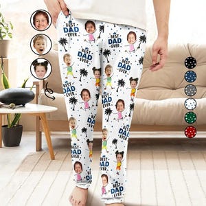 May include: White pajama trousers featuring a repeating pattern of children's faces, palm trees, and the text "BEST DAD EVER" in blue. The trousers are being held up by a person.