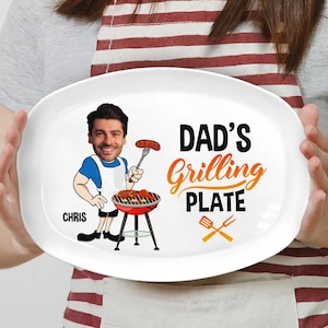 May include: White oval ceramic serving plate with a cartoon illustration of a man grilling. The text "DAD'S GRILLING PLATE" is printed in orange and black. The man's name "CHRIS" is printed in black below the illustration.