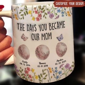Op de afbeelding: Witte keramische mok met een bloemenpatroon en de tekst "The days you became our mom". Drie manen met de namen Emily, Thomas en Sophia en hun geboortedata zijn afgebeeld.