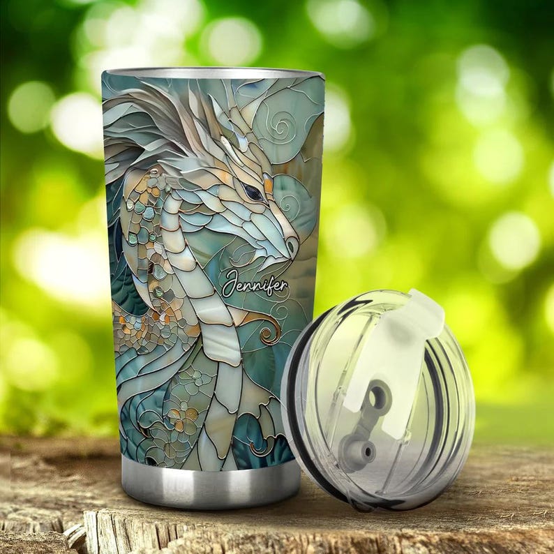 May include: A stainless steel tumbler with a lid and a colourful stained glass design of a dragon. The tumbler has the name "Jennifer" printed on it.