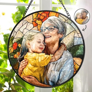May include: A stained glass window hanging with a colourful mosaic design featuring a grandmother and granddaughter hugging. The image is framed in black metal.