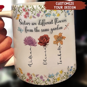 Puede incluir: Taza de cerámica blanca con un diseño floral y el texto "Sisters are different flowers from the same garden". La taza tiene tres flores con los nombres "William", "Olivia" e "Isabella" escritos debajo.