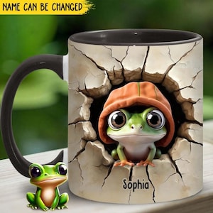 Puede incluir: Una taza de cerámica negra y blanca con una rana verde asomándose por una pared agrietada. La rana lleva una sudadera con capucha naranja y la taza dice "Sophia" en la parte inferior.