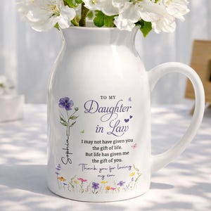 Op de afbeelding: Witte keramische kruikvaas met een handvat, gevuld met witte bloemen. De vaas is versierd met een bloemenontwerp en de tekst "To My Daughter in Law". De tekst bevat ook de naam "Sophia".