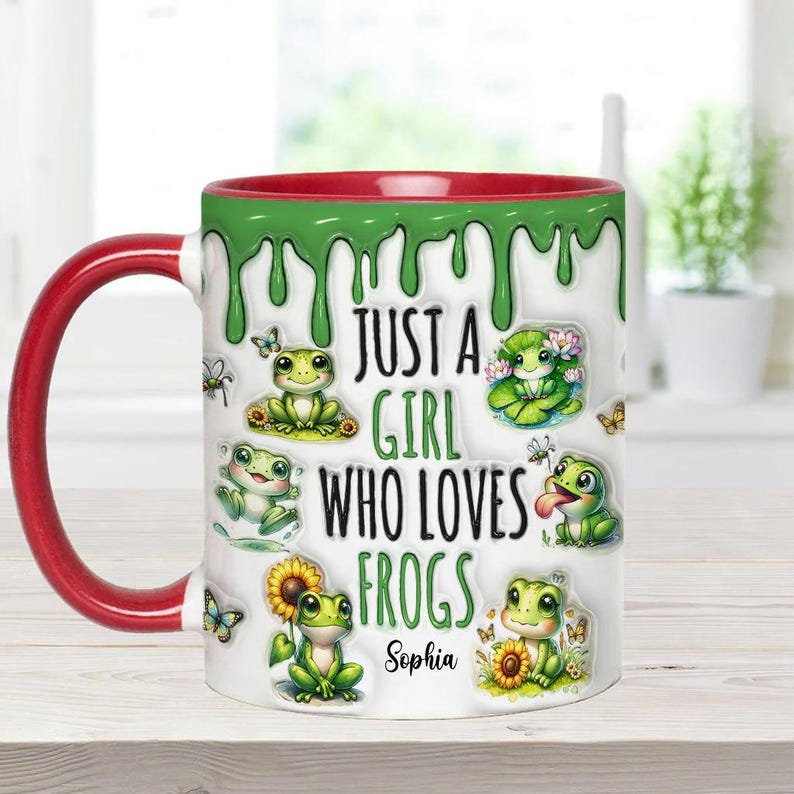 May include: A white ceramic mug with a red rim and green dripping paint design. It features a cartoon frog design and the text "Just a girl who loves frogs" with the name "Sophia" at the bottom.
