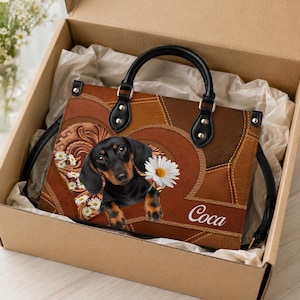 Puede incluir: Un bolso de cuero marrón con un cachorro de perro salchicha negro y fuego que sostiene una margarita blanca en la boca. El bolso tiene asas negras, un diseño de patchwork y el nombre "Coca" impreso en letra blanca en la esquina inferior.