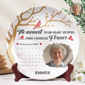 Op de afbeelding: Een wit keramiek bord met een gouden boomontwerp en twee rode kardinaals. Het bord bevat een foto van een vrouw en de tekst "The moment your heart stopped mine changed forever."  Het bord heeft ook een kalender voor februari 2025 en de naam "Jennifer".