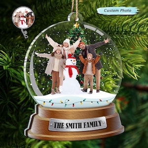 May include: A personalised Christmas bauble featuring a family of four standing in front of a decorated Christmas tree and a snowman. The bauble is a glass globe with a wooden base and the text "The Smith Family" engraved on the base.