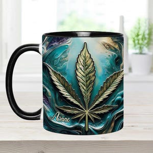 May include: A black and white coffee mug with a blue and green abstract design featuring a large marijuana leaf. The mug has a black handle and the name "Anne" is printed on the side.
