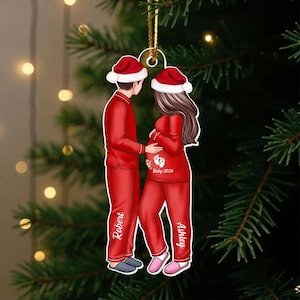 Op de afbeelding: Een kerstornament met een stel in rode pyjama's en kerstmutsen. De vrouw is zwanger, met de tekst "Baby 2026" en een voetafdrukontwerp op haar shirt. De namen "Robert" en "Ashley" staan op de pyjama.