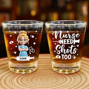 Può includere: Due bicchierini pieni di liquido ambrato. Un bicchiere presenta un'infermiera dei cartoni animati con il nome "Jasmine" e simboli medici. L'altro bicchiere ha la scritta "Nurse NEED Shots TOO" in bianco. Entrambi i bicchieri sono su una superficie di legno.