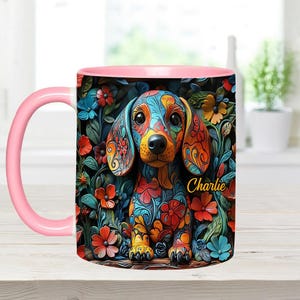 Puede incluir: Una taza de cer&aacute;mica rosa y blanca con una ilustraci&oacute;n colorida de un perro salchicha rodeado de flores. El perro est&aacute; decorado con intrincados patrones florales y el texto "Charlie" est&aacute; impreso en la taza.