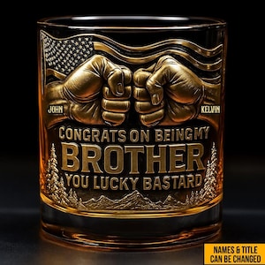 Puede incluir: Un vaso de whisky personalizado con un diseño dorado en relieve. El diseño presenta dos puños chocando, una bandera estadounidense y el texto "CONGRATS ON BEING MY BROTHER YOU LUCKY BASTARD". Se incluyen los nombres "JOHN" y "KELVIN".
