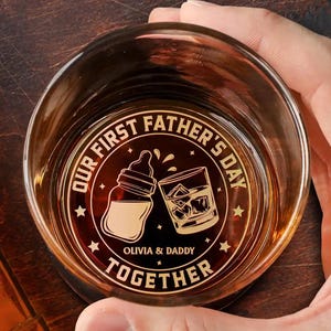 May include: A clear glass with a gold design that says "Our First Father's Day Together" with a baby bottle and a glass of whiskey. The text also includes the names "Olivia & Daddy".