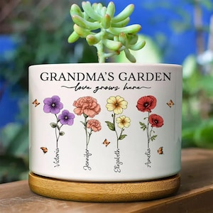 Może przedstawiać: Biała ceramiczna donica z napisem "GRANDMA'S GARDEN love grows here." Donica posiada kolorowe ilustracje kwiatowe oraz imiona "Victoria", "Jennifer", "Elizabeth" i "Amelia". Stoi na okrągłej drewnianej podstawie, z rośliną sukulentową.