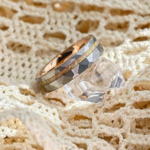 May include: A close-up of a ring with a hammered, faceted grey band and a rose gold interior. The ring features a central inlay of iridescent, speckled material. The ring rests on a clear, geometric-shaped object, set against a cream-coloured, textured background.