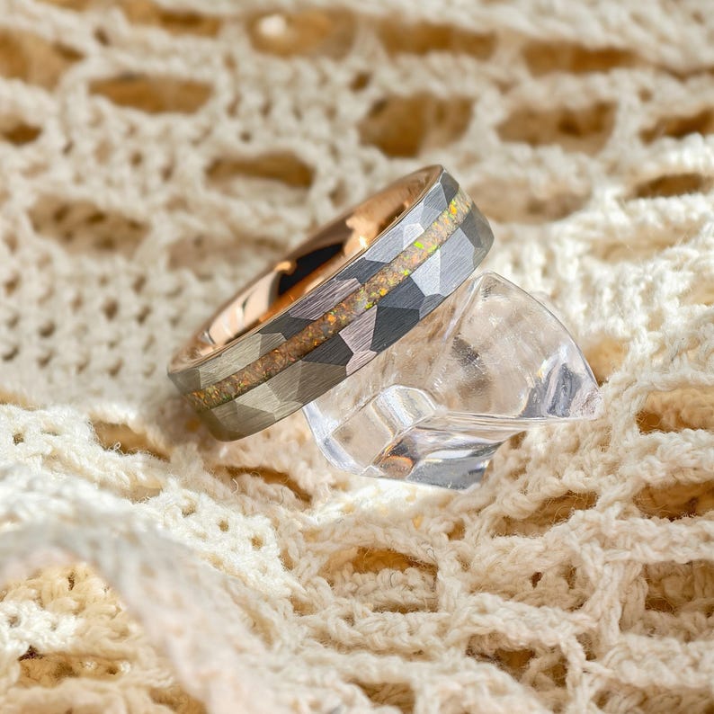 May include: A close-up of a ring with a hammered, faceted grey band and a rose gold interior. The ring features a central inlay of iridescent, speckled material. The ring rests on a clear, geometric-shaped object, set against a cream-coloured, textured background.