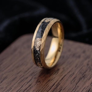 Peut inclure: Une bague dorée avec un bord extérieur facetté et une incrustation centrale en matériau noir parsemé de paillettes dorées. L'anneau présente une finition polie sur les côtés et un canal central texturé, sombre et moucheté.