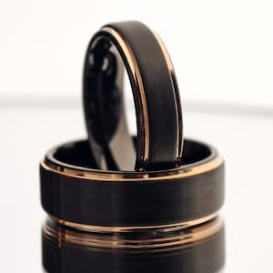 May include: Two black rings with a brushed metal finish and rose gold edges. One ring is standing upright inside the other, creating a layered effect. The rings have a modern, minimalist design.