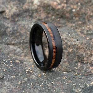 Könnte beinhalten: Ein schwarzer Ring aus Wolframcarbid mit einer braunen Holzeinlage. Der Ring hat eine polierte Oberfläche und eine abgeschrägte Kante.