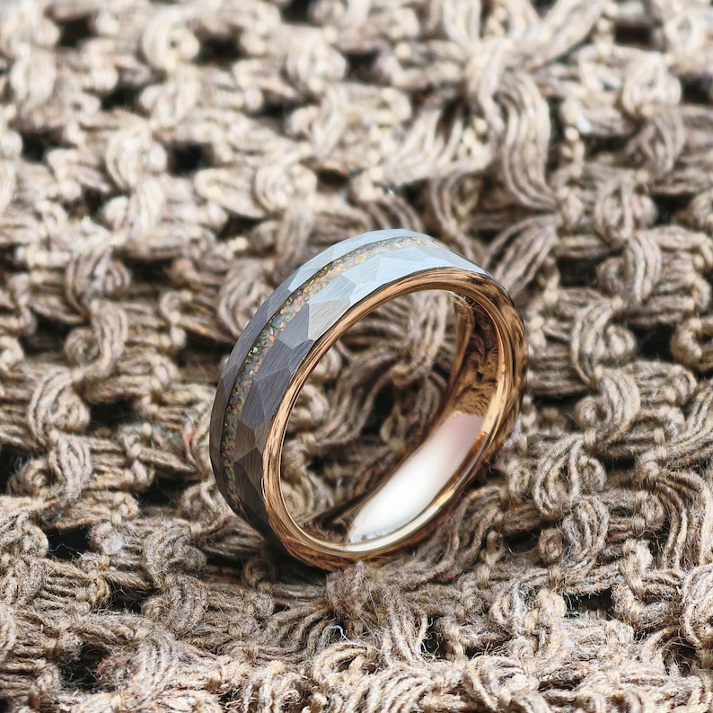 May include: A rose gold wedding band with a faceted grey exterior and a central inlay of iridescent material. The ring is set against a textured, brown background, highlighting its metallic sheen and intricate design.