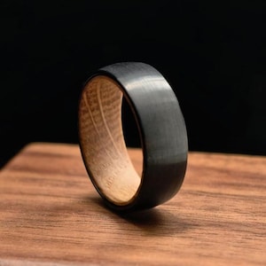 Puede incluir: Un anillo negro con interior de madera. El anillo tiene un exterior negro mate liso y un interior de madera clara y texturizada. El anillo se muestra sobre una superficie de madera sobre un fondo oscuro.
