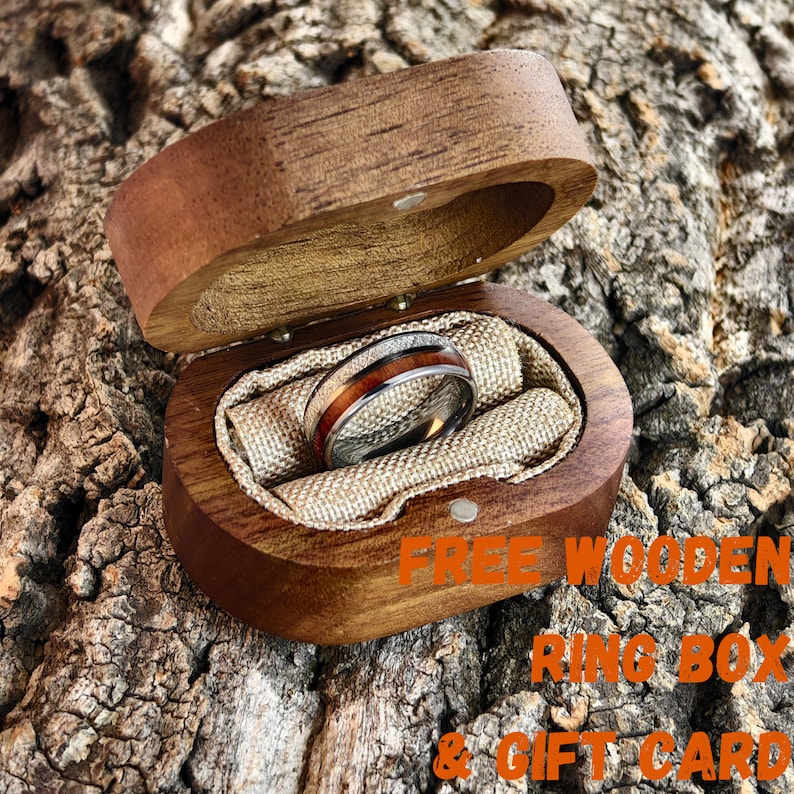 May include: A silver ring with a wood inlay is displayed inside an open, oval-shaped wooden ring box. The ring box is lined with beige fabric and rests on a textured, brown surface. The text "FREE WOODEN RING BOX & GIFT CARD" is visible.