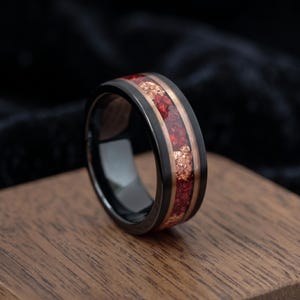 Puede incluir: Un anillo de banda negra con una incrustación central de piedra roja triturada y escamas de cobre metálico. El anillo tiene un acabado liso y pulido en los bordes exteriores y una superficie interior oscura y reflectante, mostrando un contraste marcado entre el metal oscuro y la incrustación texturizada.