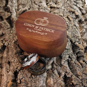 May include: A wooden ring box engraved with "CINDY & PATRICK" and the date "11.11.2024", with two wedding rings. Two rings are displayed in front of the box. One ring has a wood inlay and the other is black.