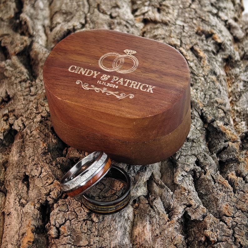 May include: A wooden ring box engraved with "CINDY & PATRICK" and the date "11.11.2024", with two wedding rings. Two rings are displayed in front of the box. One ring has a wood inlay and the other is black.