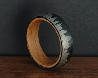 May include: A unique ring with a dark gray exterior featuring a silver tree line design. The interior of the ring is a light brown wood. The ring is displayed on a wooden surface.