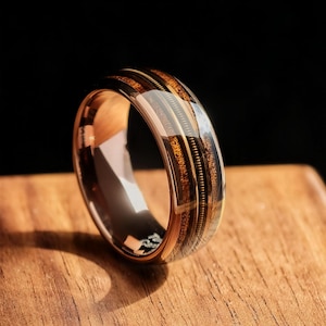 Peut inclure: Bague en or rose avec un motif incrusté de grain de bois et de métal. La bague a une finition polie et est posée sur une surface en bois. L'incrustation présente une combinaison de tons bruns foncés et clairs avec un accent métallique.