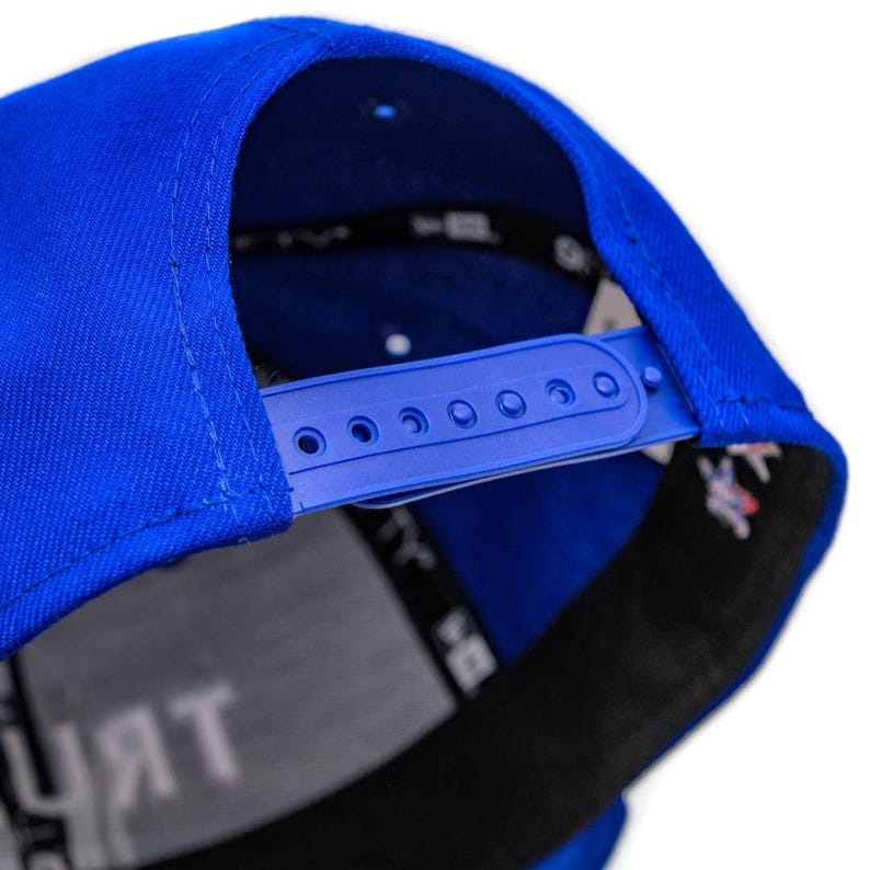 Puede incluir: Una gorra de b&eacute;isbol azul real con una correa ajustable negra y una fila de agujeros para ajustar el tama&ntilde;o. La gorra tiene un lado inferior negro y gris con la palabra "TRUST" impresa en blanco.