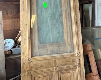 May include: Antique wooden door with a glass panel at the top and two wooden panels at the bottom. The door has decorative carvings and a keyhole. The wood has a natural, aged appearance. The door is leaning against other architectural elements.